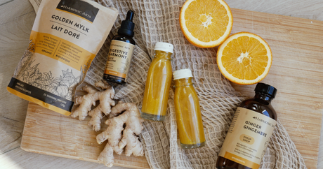 Image features two Digestive Wellness shots in a flat lay next to Harmonic Arts Golden Mylk Herbal Latte, Digestive Harmony Tincture Blend and Ginger Syrup. The wellness shots are also surrounded by two pieces of ginger and two orange slices.