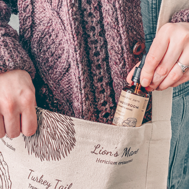 Image features a person wearing jeans and a mauve knit cardigan placing a Harmonic Arts Tincture Blend into a mushroom tote bag.